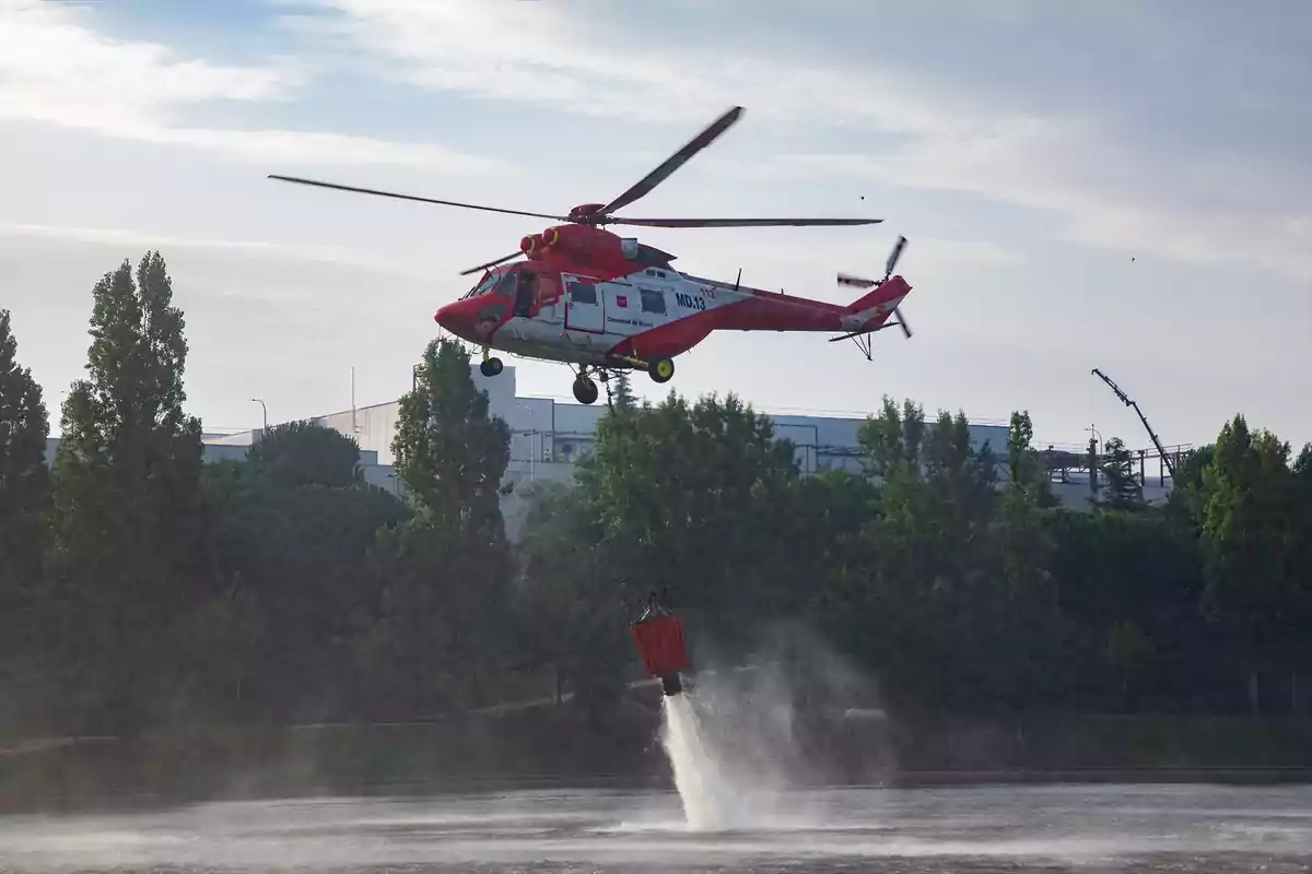 Helicóptero rojo recogiendo agua de un lago con un cubo especial rodeado de árboles y edificios al fondo Helicóptero rojo recogiendo agua de un lago con un cubo especial rodeado de árboles y edificios al fondo