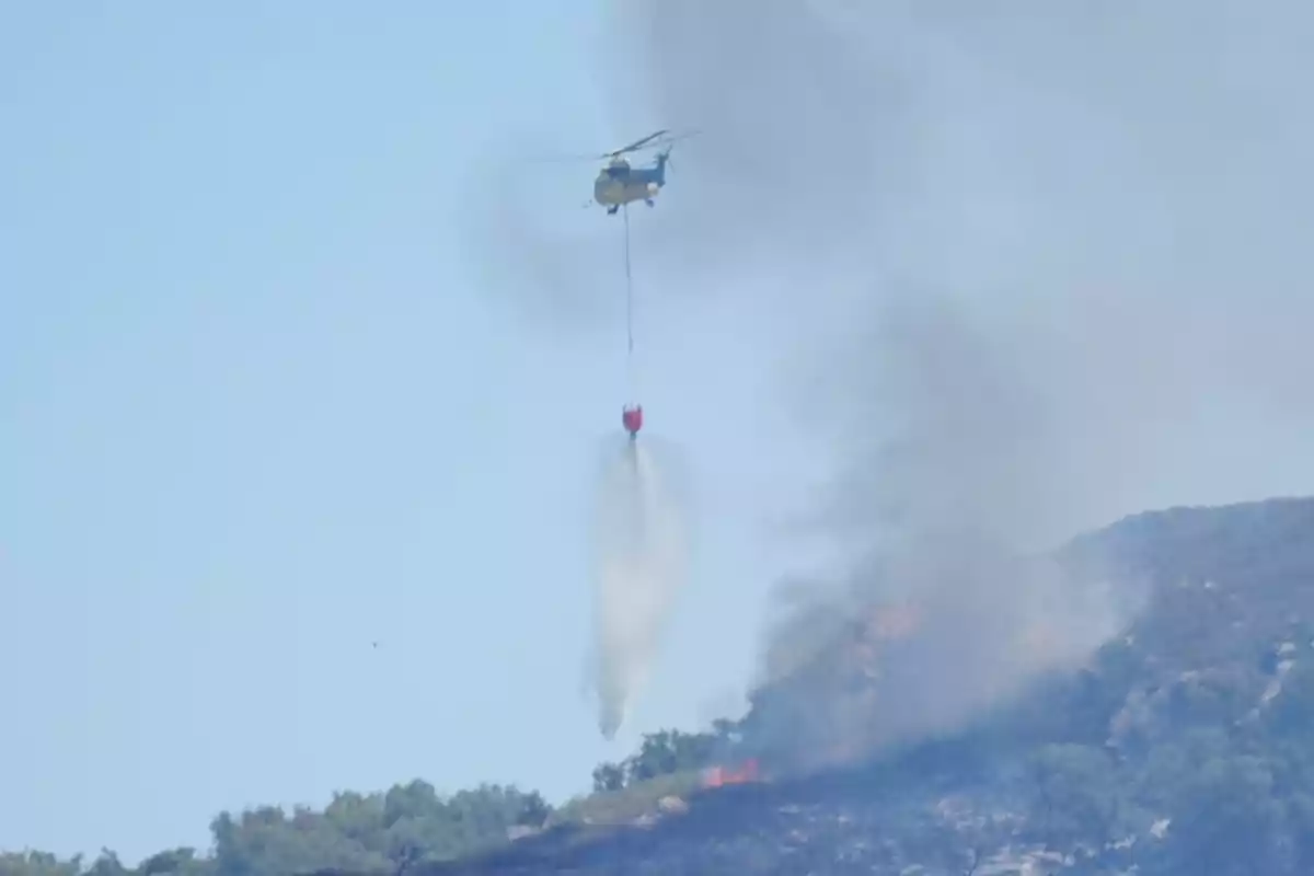 Helicóptero arrojando agua sobre un incendio forestal en una colina mientras se eleva humo al cielo