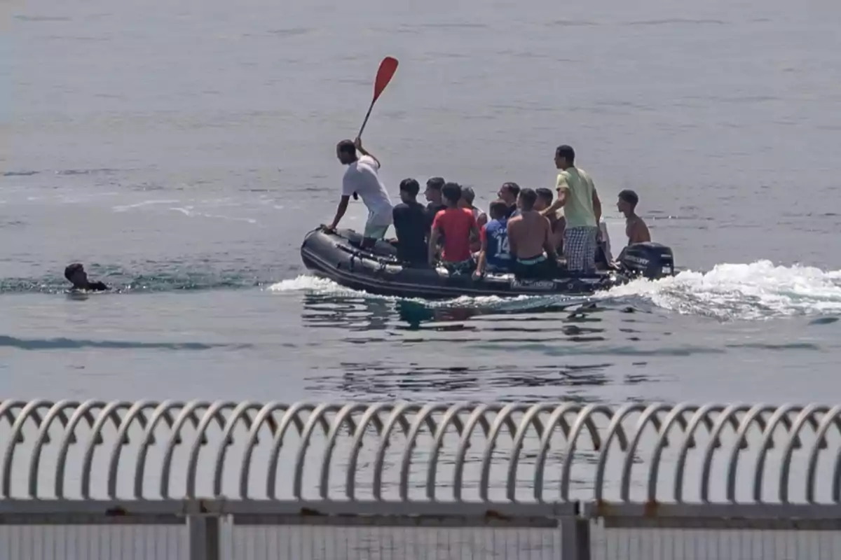 Un grupo de personas viaja en una lancha inflable sobre el mar mientras una persona nada cerca de la embarcación y en primer plano se observa una valla metálica desenfocada.