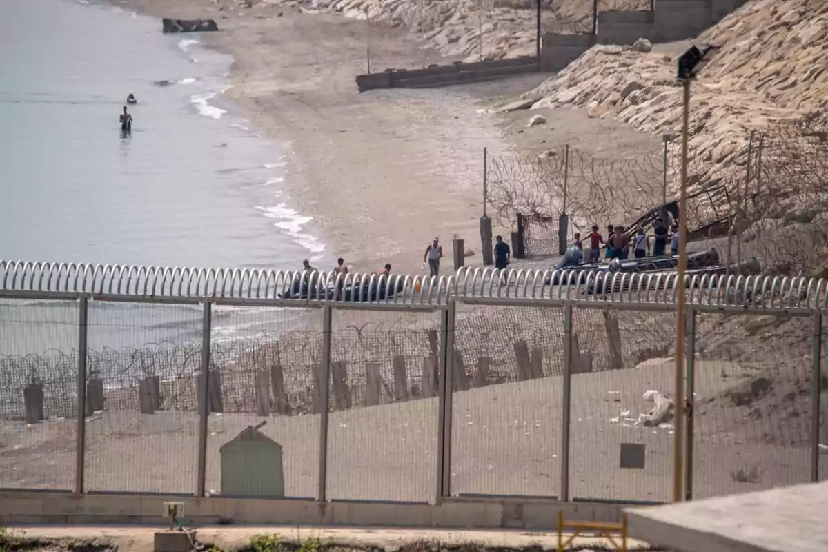 Personas en la playa junto a una valla fronteriza con alambre de púas y varias personas en el agua acercándose a la orilla Personas en la playa junto a una valla fronteriza con alambre de púas y varias personas en el agua acercándose a la orilla