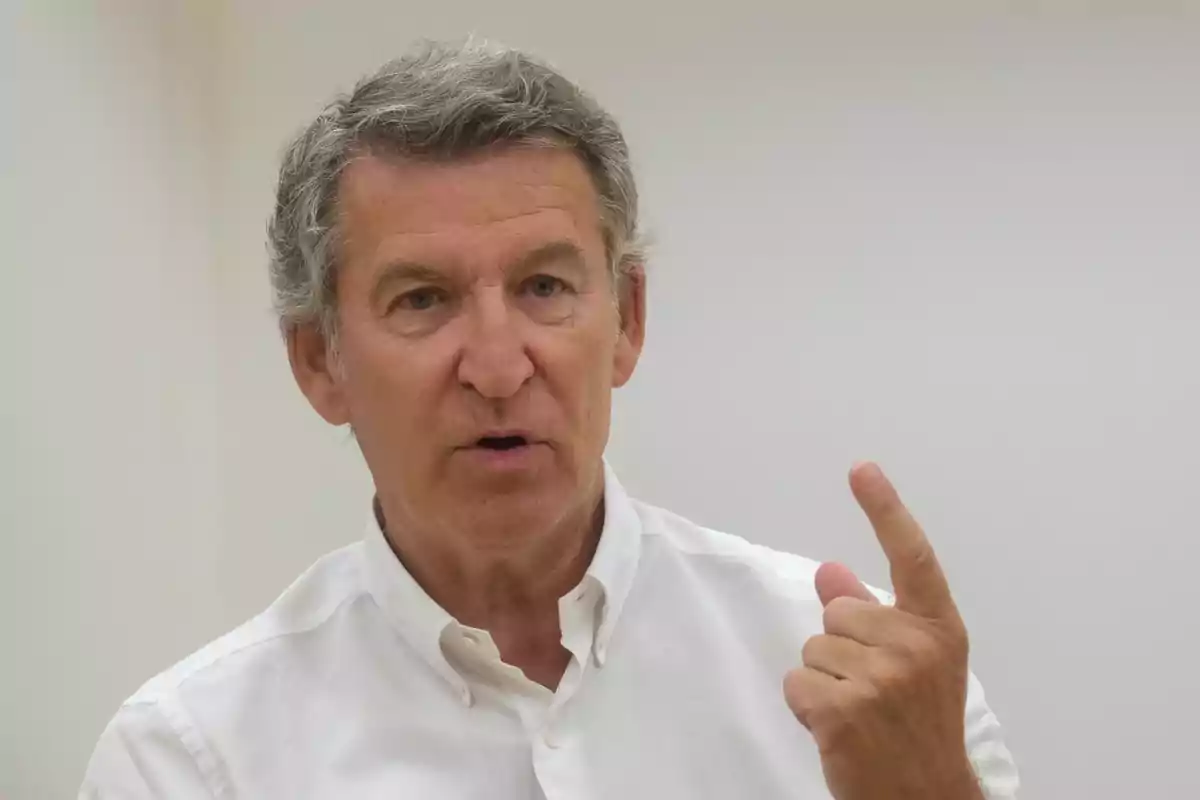 Hombre de cabello canoso con camisa blanca levantando el dedo índice mientras habla en un entorno interior iluminado