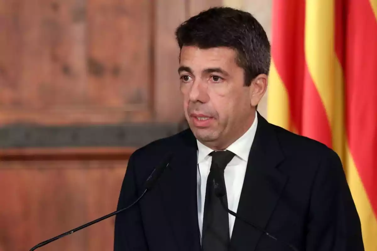 Hombre de cabello corto y oscuro con traje y corbata negra hablando frente a dos micrófonos con una bandera de fondo