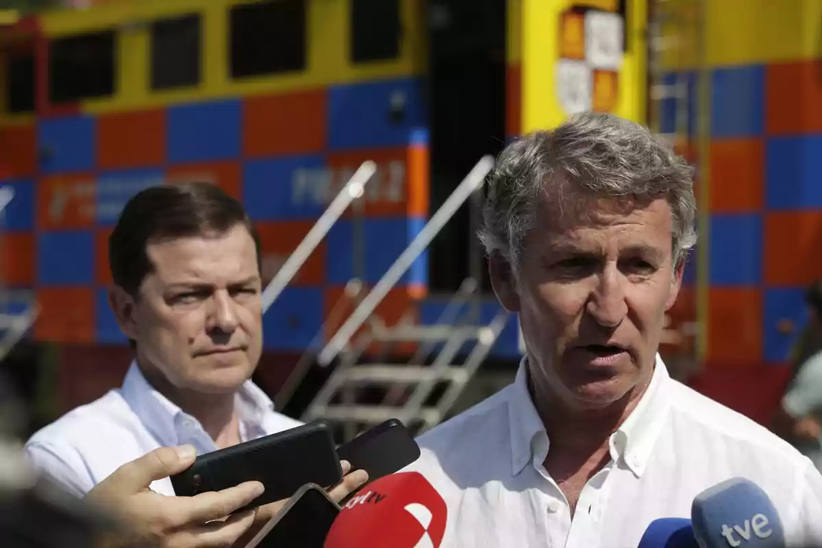 Dos hombres con camisas blancas responden preguntas de la prensa frente a un vehículo de colores llamativos con micrófonos de diferentes medios en primer plano.