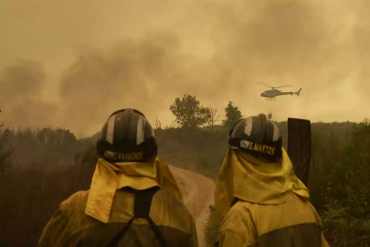 Dos bomberos observan un helicóptero que combate un incendio forestal en medio de una densa nube de humo