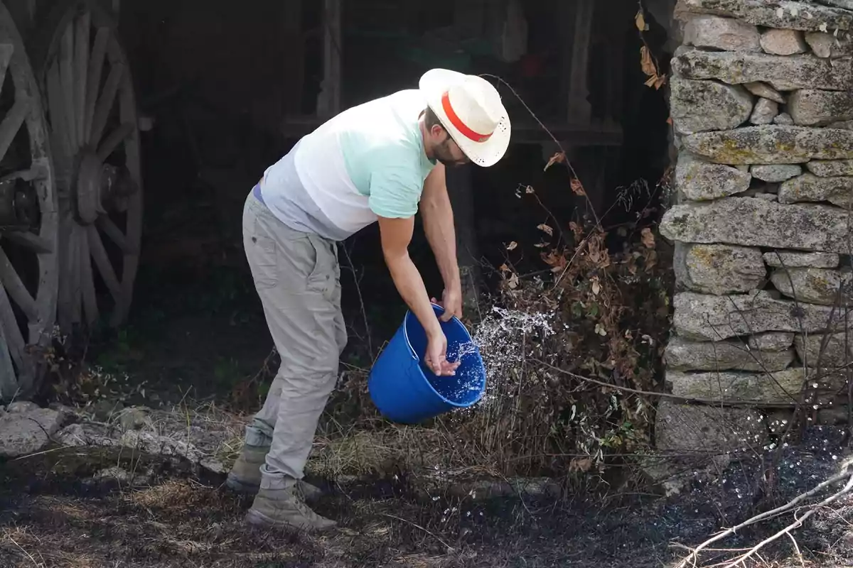 Hombre con sombrero vertiendo agua de un balde azul sobre el suelo seco junto a una pared de piedra y una carreta antigua