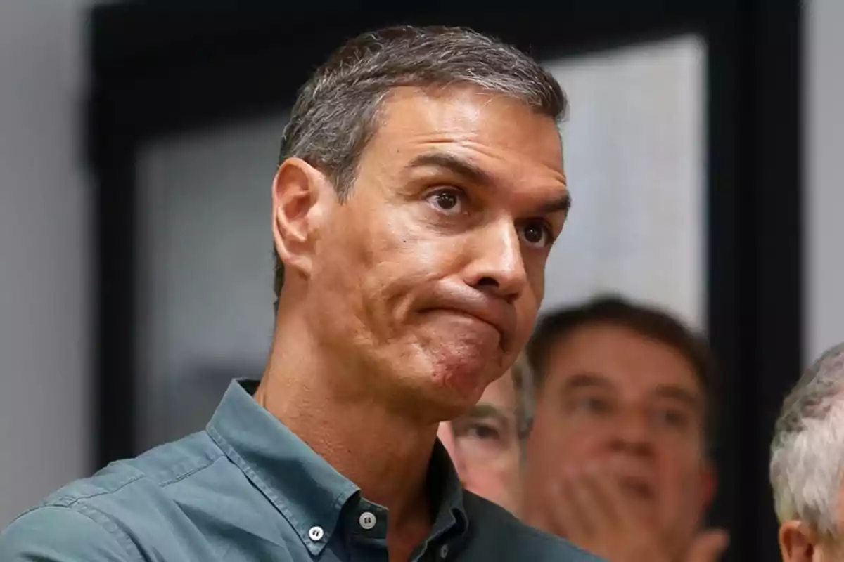 Hombre de cabello corto y canoso con expresión seria y labios apretados, vistiendo camisa verde, en un ambiente interior con personas al fondo desenfocadas