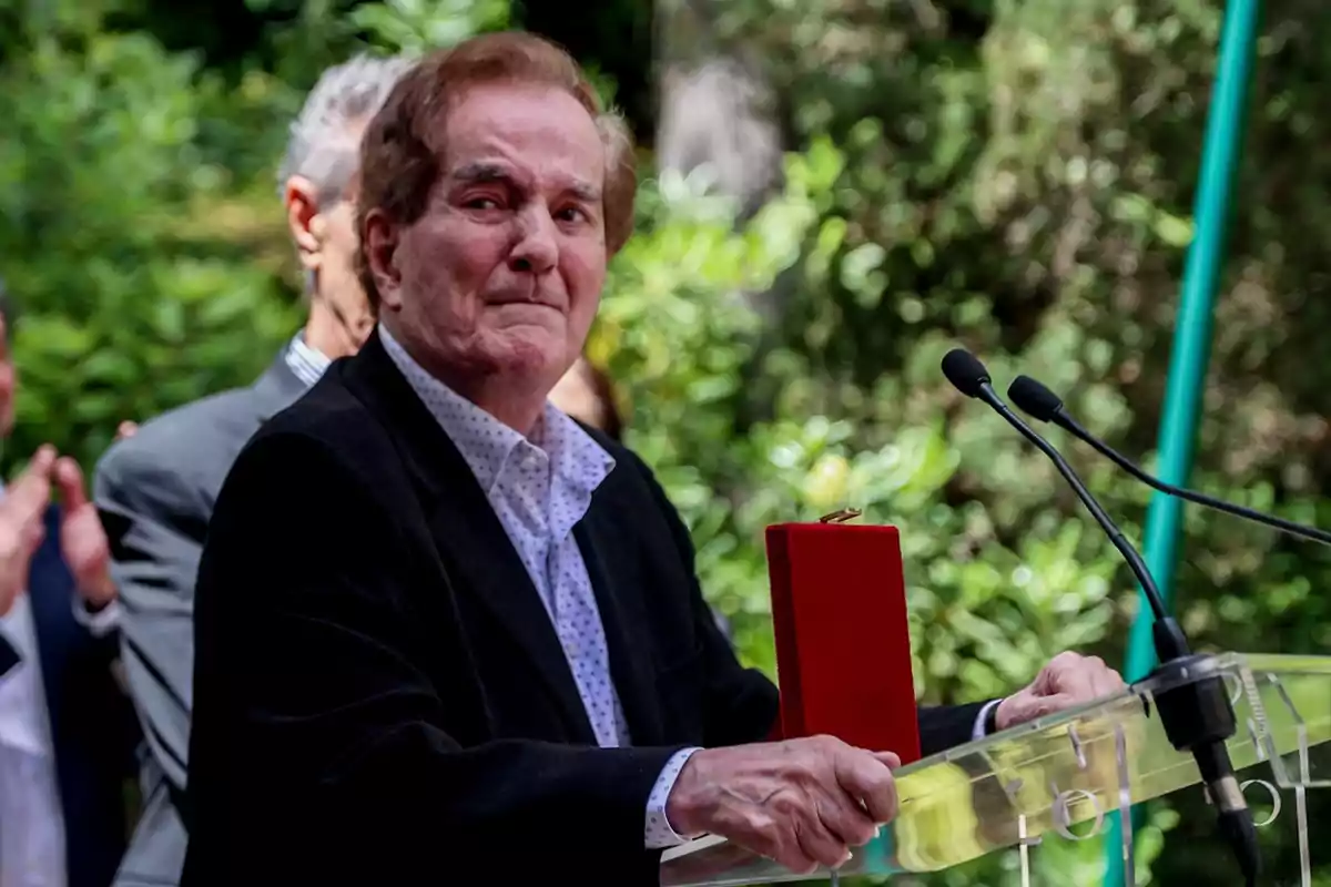 Hombre mayor de pie frente a un atril con micrófonos sosteniendo una caja roja mientras personas al fondo aplauden en un entorno al aire libre con vegetación