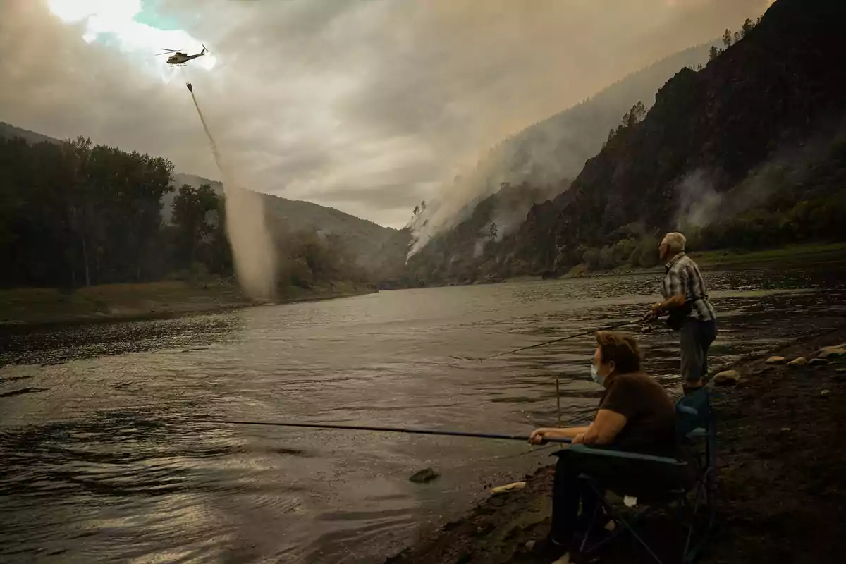 Dos personas pescando en la orilla de un río mientras un helicóptero arroja agua sobre un incendio forestal en las montañas al fondo Dos personas pescando en la orilla de un río mientras un helicóptero arroja agua sobre un incendio forestal en las montañas al fondo