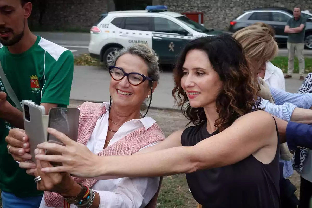 Dos mujeres sonríen mientras se toman una selfie con un teléfono móvil en un ambiente al aire libre, al fondo se observa un coche de la Guardia Civil y otras personas
