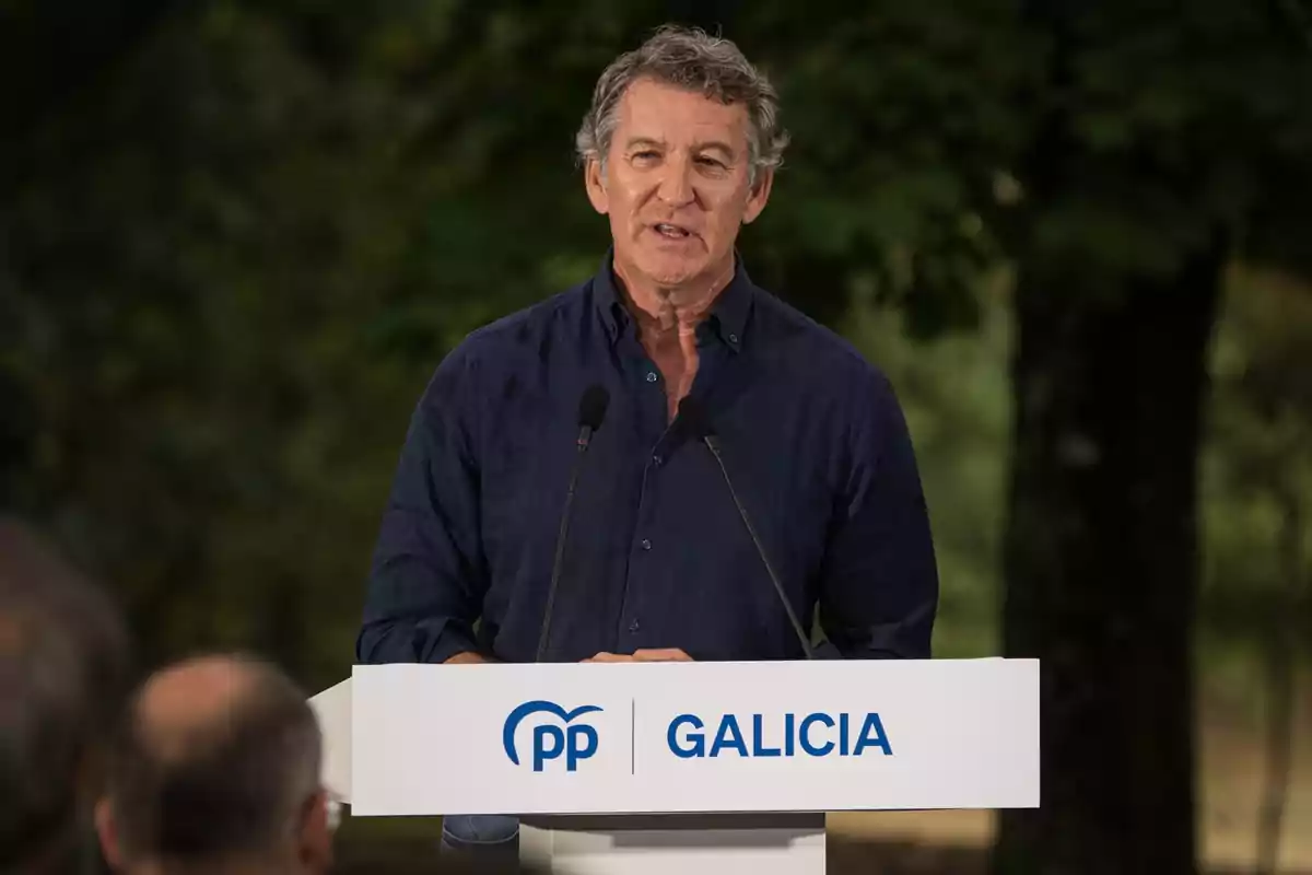 Hombre de cabello canoso hablando en un atril con el logo de PP y la palabra Galicia en un entorno al aire libre