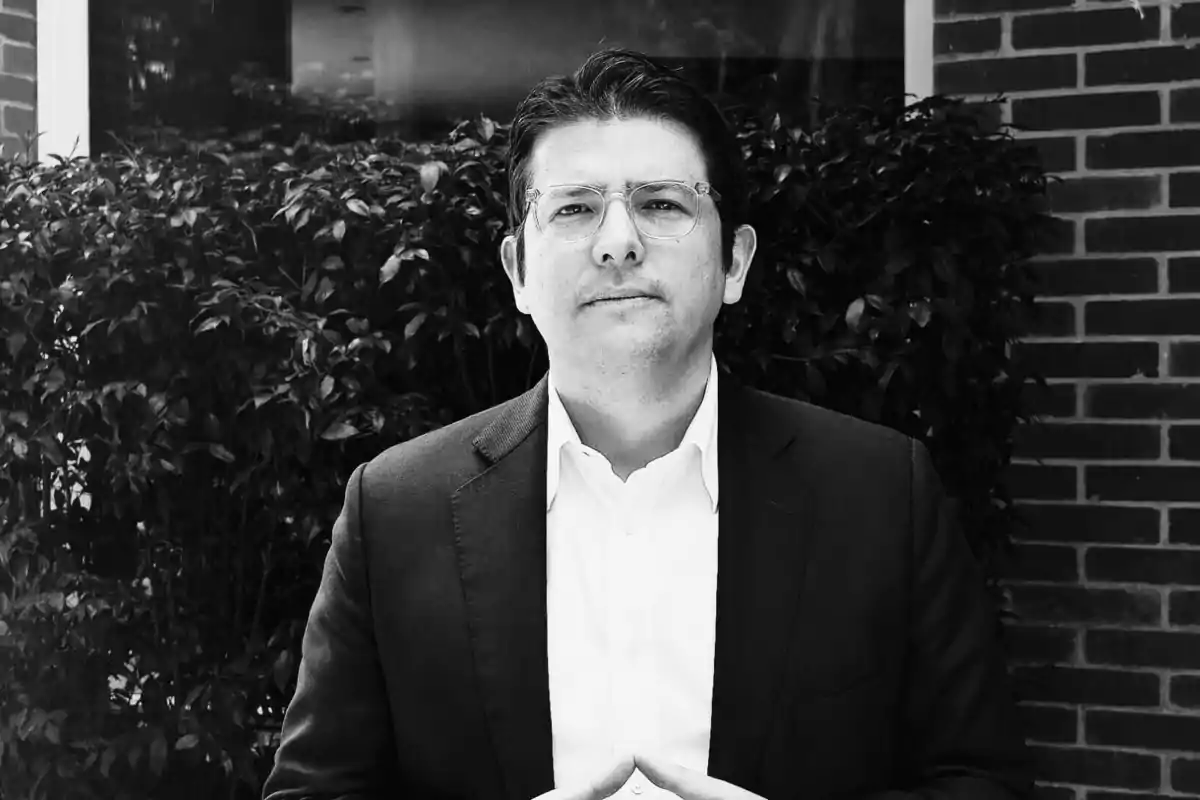 Hombre con gafas y traje oscuro posando frente a un arbusto y una pared de ladrillos en una imagen en blanco y negro