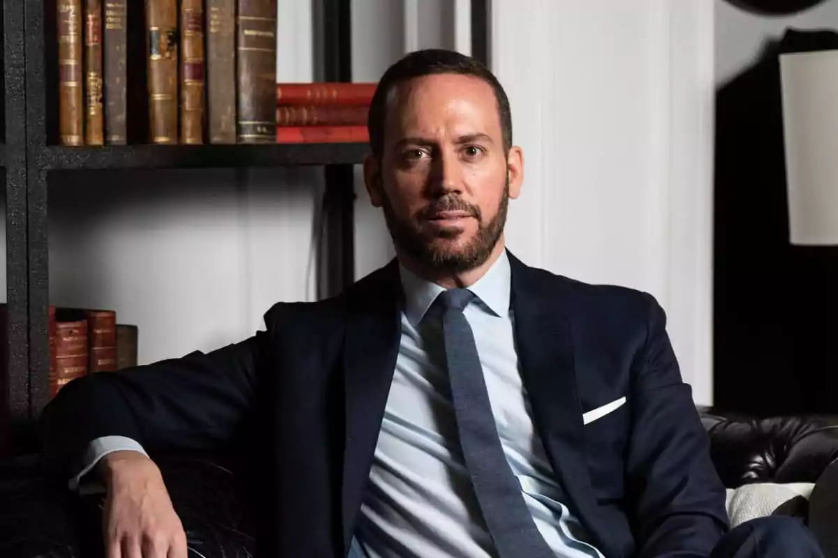Hombre con barba y traje azul marino sentado en un sofá de cuero negro con estantería de libros antiguos al fondo Hombre con barba y traje azul marino sentado en un sofá de cuero negro con estantería de libros antiguos al fondo