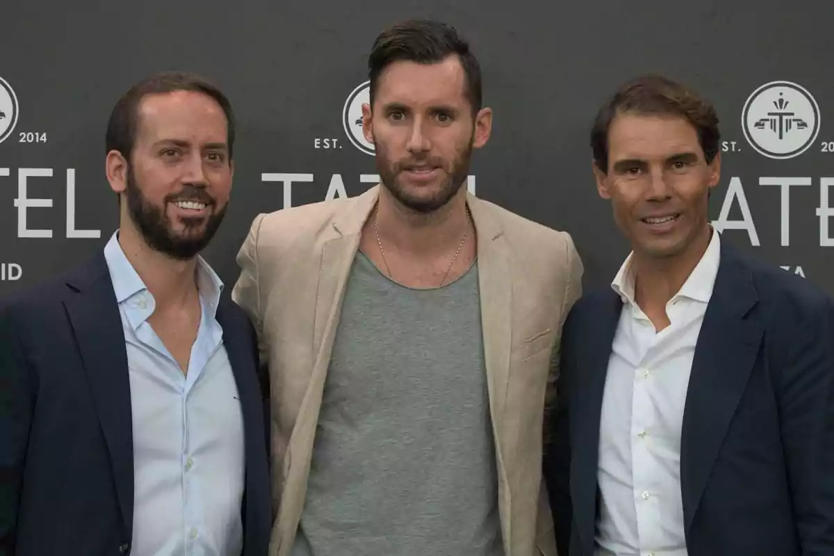 Tres hombres posan juntos frente a un fondo gris con logotipos y letras blancas, todos visten ropa formal y sonríen a la cámara. Tres hombres posan juntos frente a un fondo gris con logotipos y letras blancas, todos visten ropa formal y sonríen a la cámara.