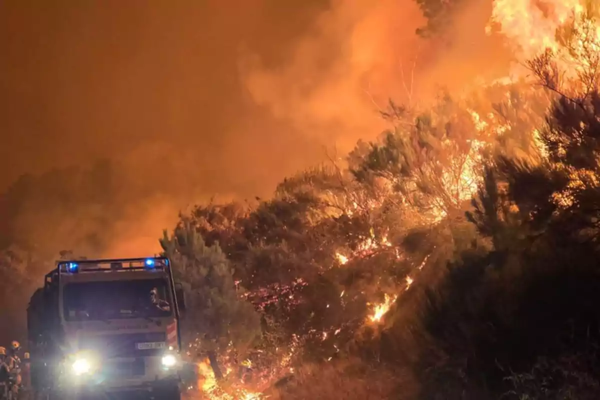 Un camión de bomberos se encuentra junto a un incendio forestal de gran magnitud con llamas y humo cubriendo el cielo Un camión de bomberos se encuentra junto a un incendio forestal de gran magnitud con llamas y humo cubriendo el cielo