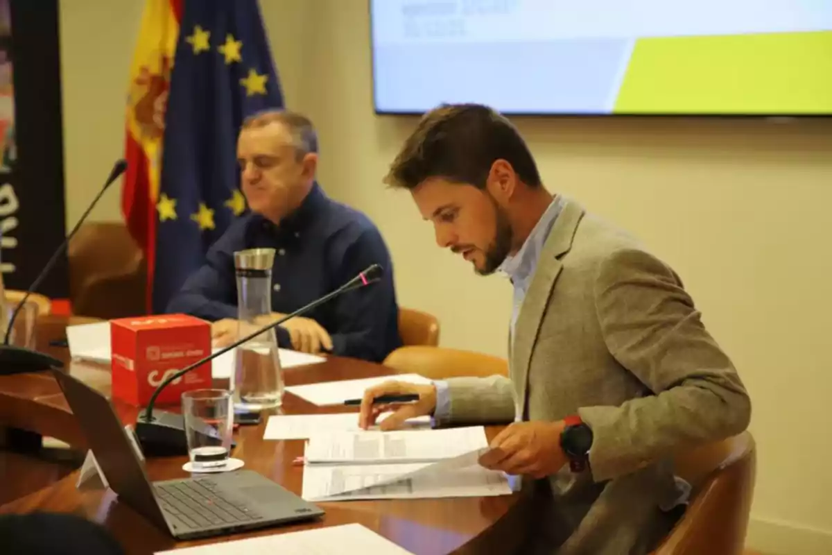 Dos hombres sentados en una mesa de reuniones revisan documentos frente a una computadora portátil con banderas de España y la Unión Europea al fondo Dos hombres sentados en una mesa de reuniones revisan documentos frente a una computadora portátil con banderas de España y la Unión Europea al fondo