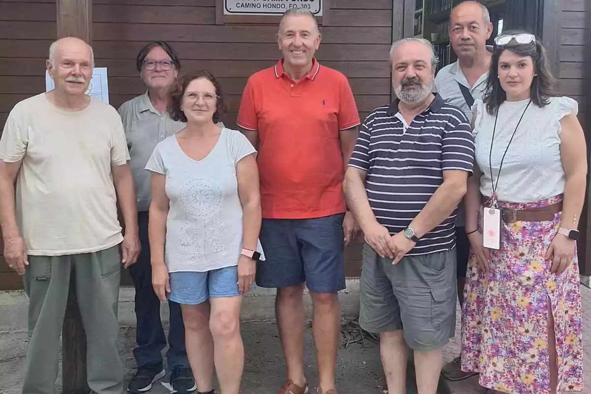 Siete personas posan juntas al aire libre frente a una pared de madera, algunas sonríen y visten ropa casual de verano.