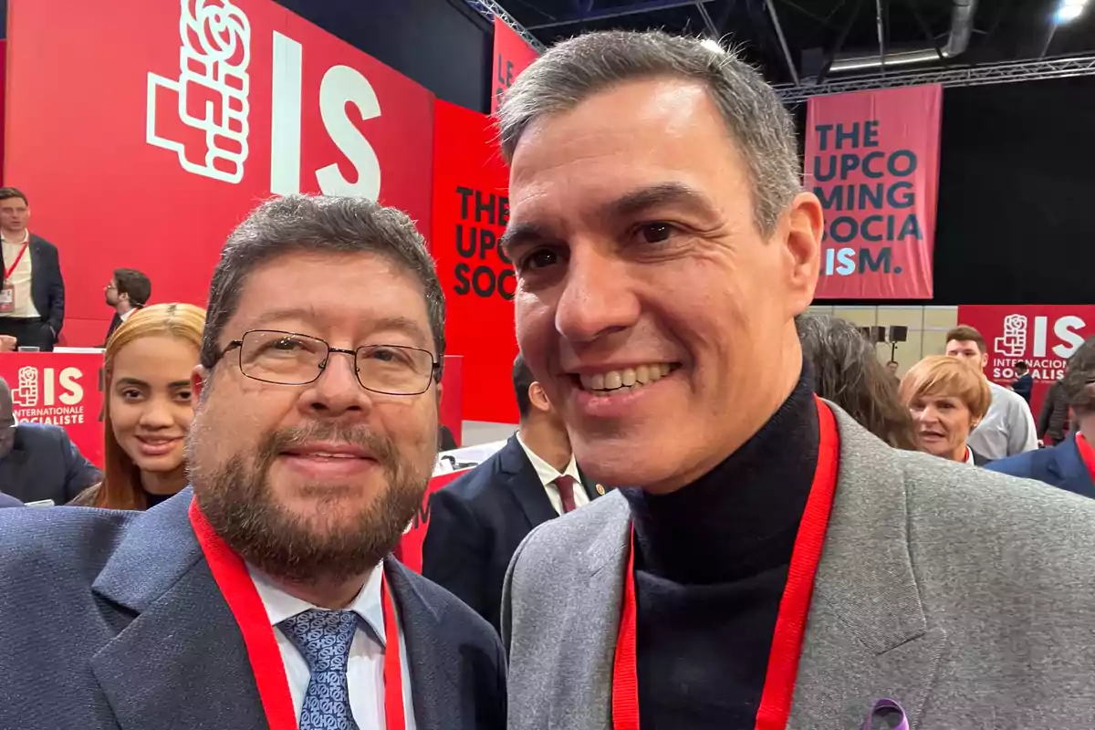 Dos hombres sonríen para una selfie en un evento con fondo rojo y letreros de Internacional Socialista rodeados de otras personas asistentes