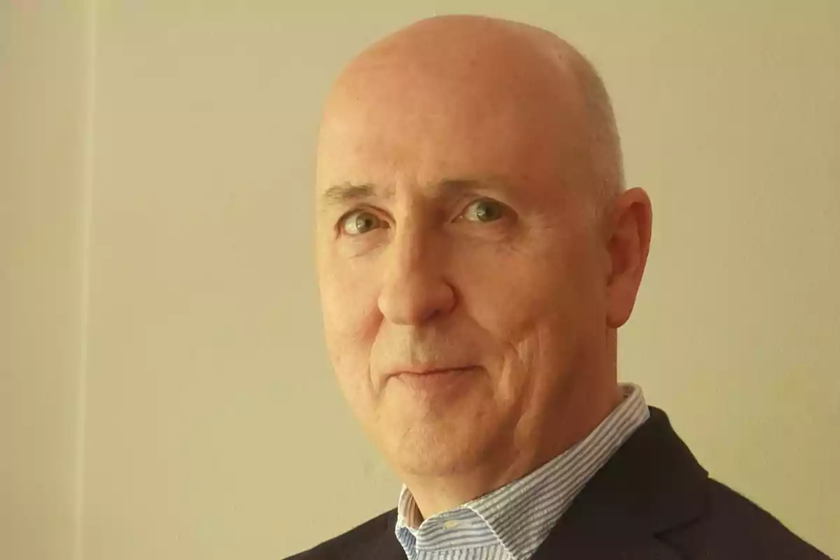Hombre calvo de mediana edad con camisa azul y saco oscuro mirando a la cámara frente a una pared clara
