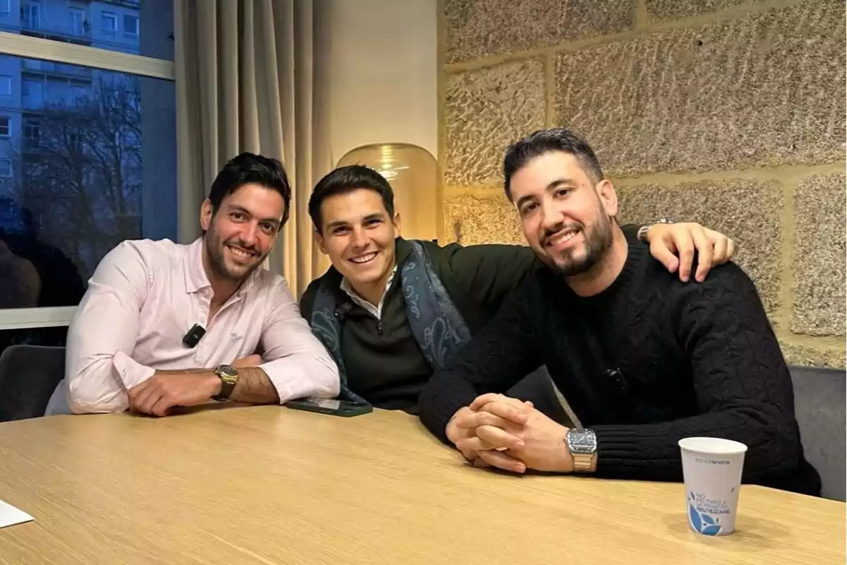 Tres hombres sonríen sentados alrededor de una mesa de madera en una sala iluminada, con una pared de piedra al fondo y una ventana a la izquierda.