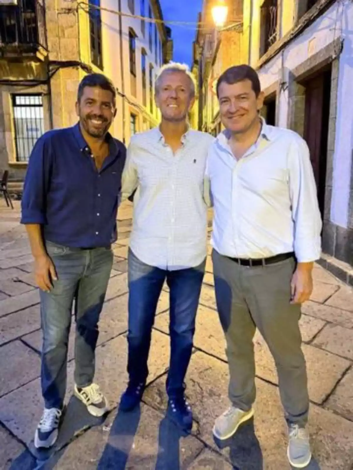 Tres hombres sonríen y posan juntos en una calle empedrada al atardecer con edificios antiguos a su alrededor Tres hombres sonríen y posan juntos en una calle empedrada al atardecer con edificios antiguos a su alrededor