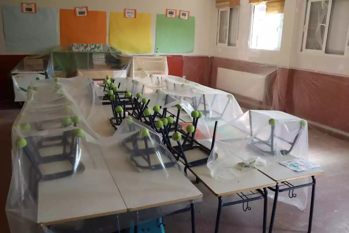 Aula escolar con mesas y sillas cubiertas por plásticos protectores y paredes decoradas con cartulinas de colores