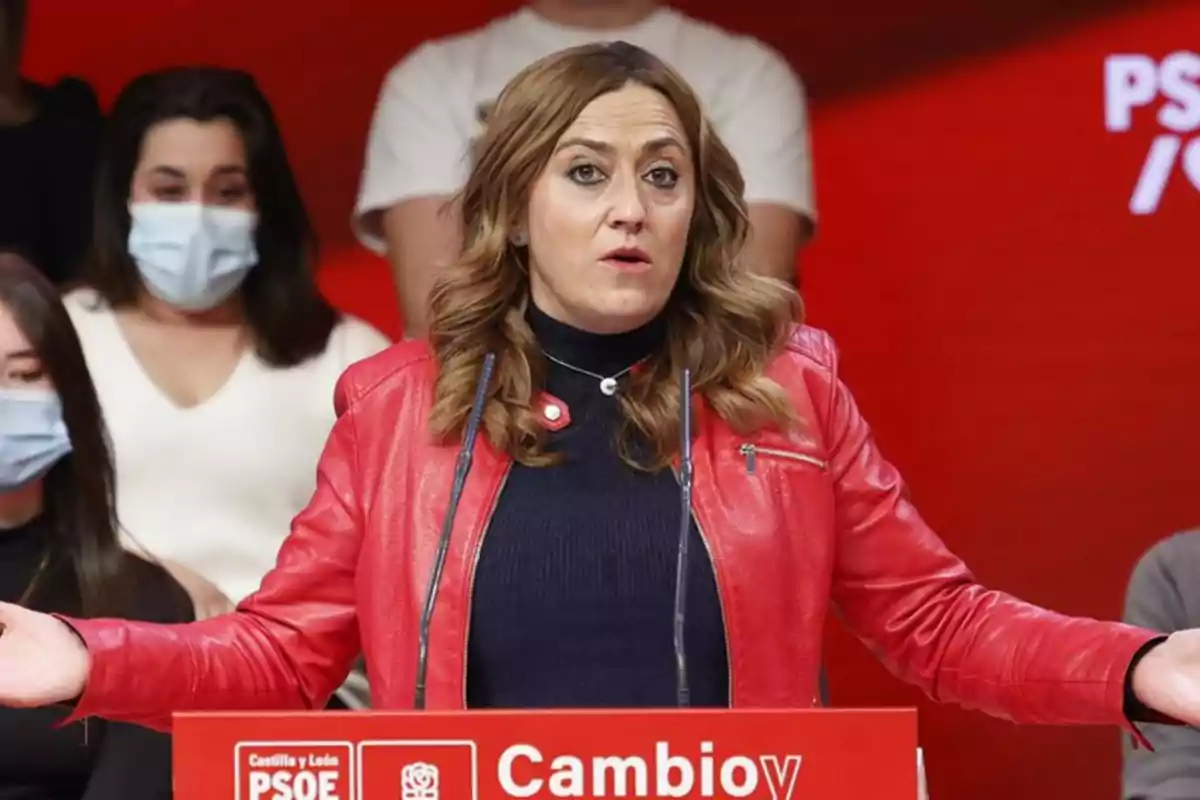 Mujer con chaqueta roja hablando en un atril durante un evento del PSOE con personas al fondo usando mascarillas Mujer con chaqueta roja hablando en un atril durante un evento del PSOE con personas al fondo usando mascarillas