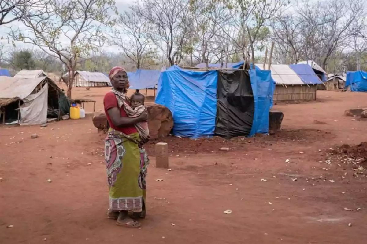 Mujer con un bebé en brazos de pie en un campamento con refugios improvisados hechos de lonas y ramas en un terreno de tierra roja