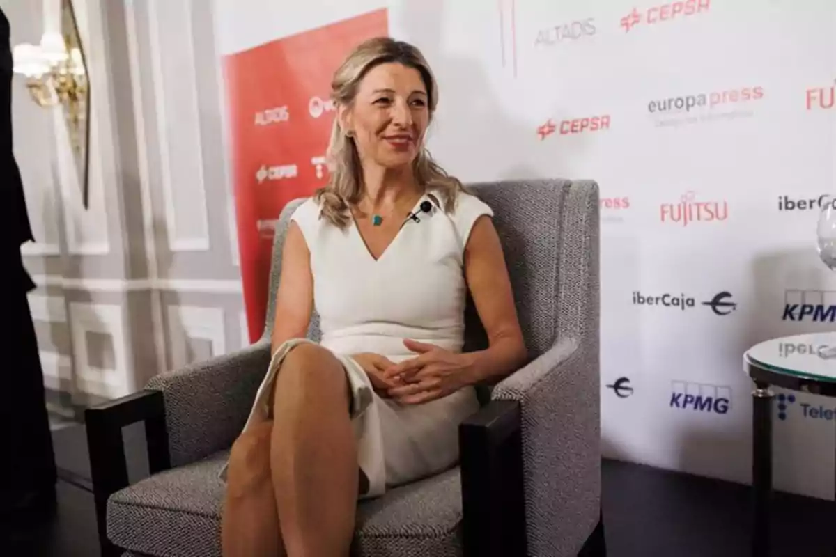 Mujer sentada en un sillón gris durante un evento, viste un vestido blanco y lleva un micrófono de solapa, al fondo se ven logotipos de empresas en un panel blanco y rojo. Mujer sentada en un sillón gris durante un evento, viste un vestido blanco y lleva un micrófono de solapa, al fondo se ven logotipos de empresas en un panel blanco y rojo.