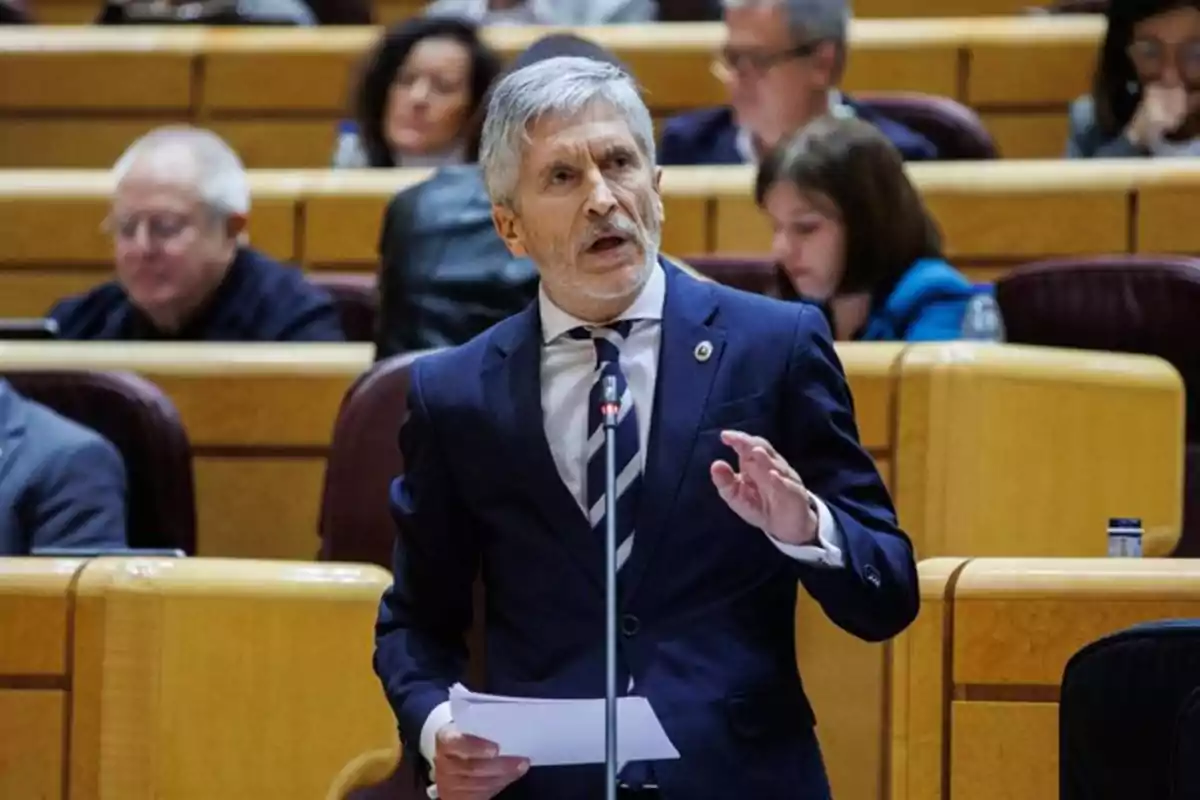 Hombre de cabello canoso y barba hablando en un micrófono mientras sostiene papeles en una sala de conferencias con varias personas sentadas al fondo Hombre de cabello canoso y barba hablando en un micrófono mientras sostiene papeles en una sala de conferencias con varias personas sentadas al fondo