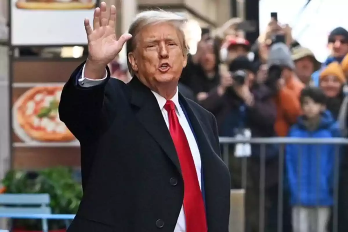 Hombre de cabello rubio con traje oscuro y corbata roja saludando con la mano levantada mientras camina por la calle frente a un grupo de personas Hombre de cabello rubio con traje oscuro y corbata roja saludando con la mano levantada mientras camina por la calle frente a un grupo de personas