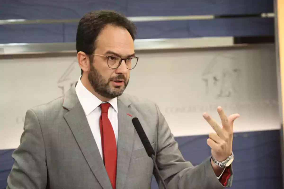 Hombre con gafas y barba usando traje gris y corbata roja hablando frente a un micrófono y levantando tres dedos de la mano derecha Hombre con gafas y barba usando traje gris y corbata roja hablando frente a un micrófono y levantando tres dedos de la mano derecha