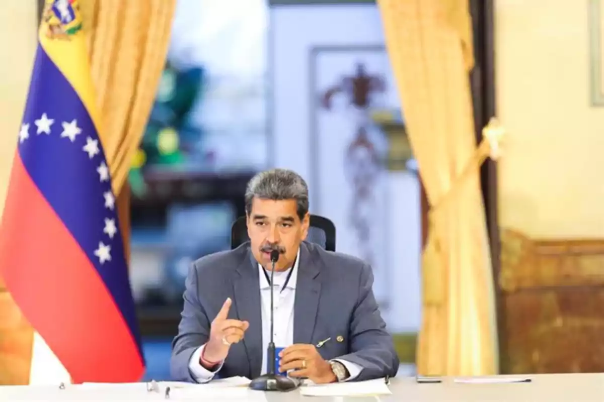 Hombre de cabello canoso y bigote hablando en una conferencia con micrófono frente a una bandera de Venezuela y cortinas amarillas Hombre de cabello canoso y bigote hablando en una conferencia con micrófono frente a una bandera de Venezuela y cortinas amarillas