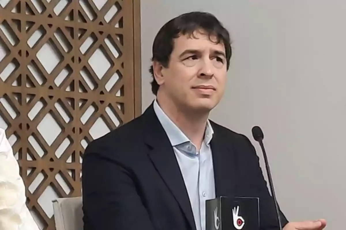Hombre de cabello oscuro con saco azul y camisa clara sentado frente a un micrófono en una sala con decoración de madera geométrica Hombre de cabello oscuro con saco azul y camisa clara sentado frente a un micrófono en una sala con decoración de madera geométrica