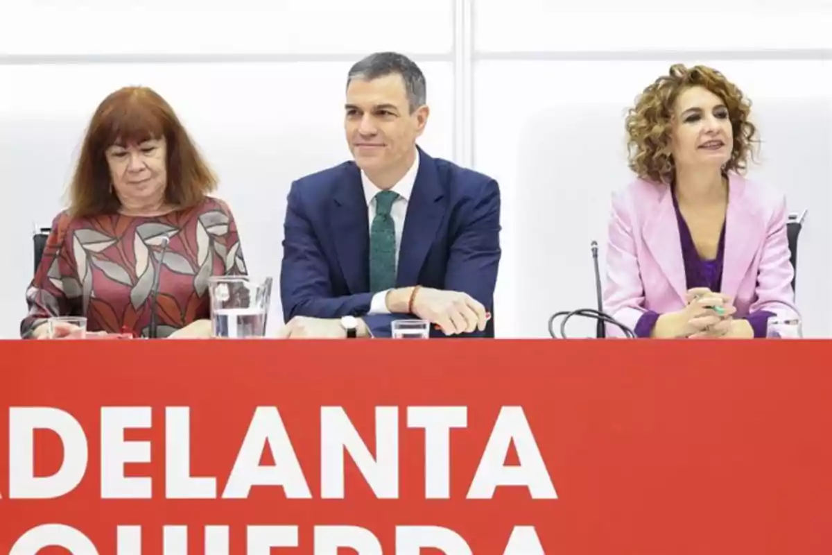 Tres personas sentadas en una mesa con un cartel rojo al frente que tiene letras blancas grandes Tres personas sentadas en una mesa con un cartel rojo al frente que tiene letras blancas grandes