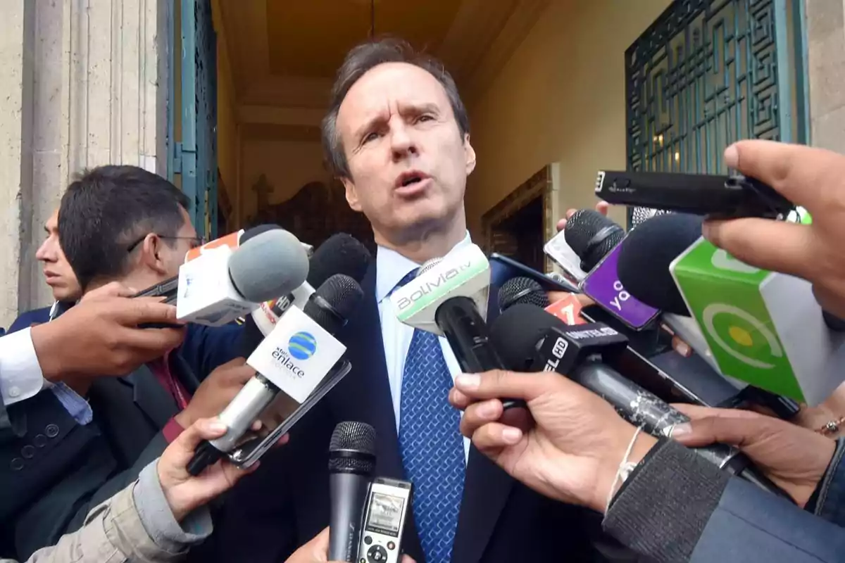 Un hombre de traje y corbata es rodeado por varios micrófonos y reporteros mientras da una declaración a la prensa en la entrada de un edificio Un hombre de traje y corbata es rodeado por varios micrófonos y reporteros mientras da una declaración a la prensa en la entrada de un edificio