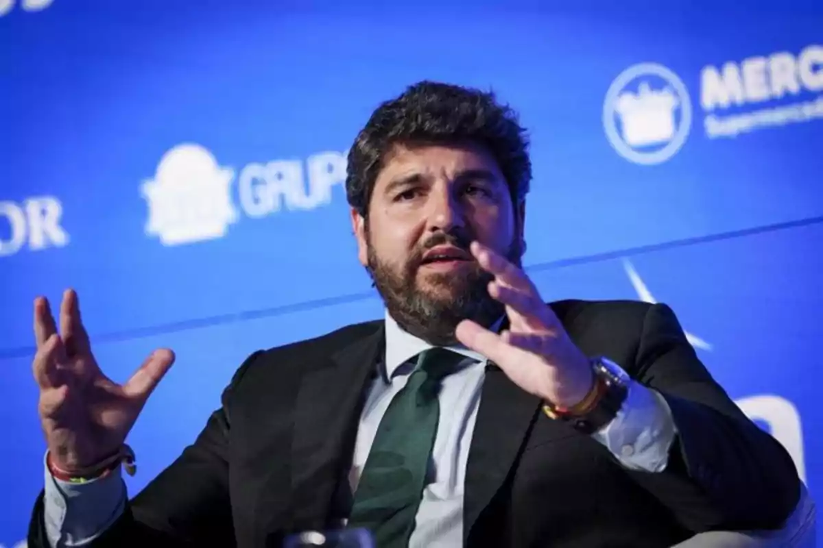 Hombre con barba y traje oscuro gesticulando mientras habla en un evento con fondo azul y logotipos desenfocados. Hombre con barba y traje oscuro gesticulando mientras habla en un evento con fondo azul y logotipos desenfocados.