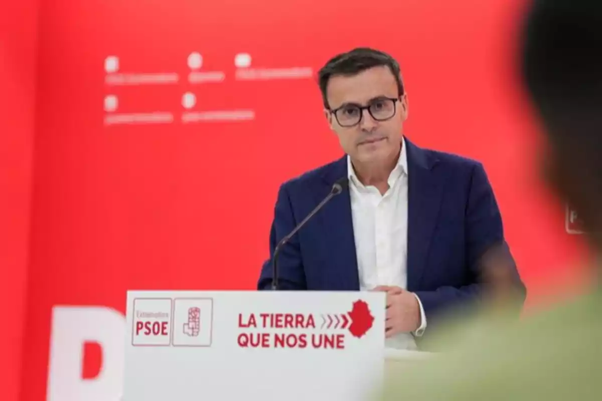 Hombre con gafas y traje azul hablando en un atril con fondo rojo y logotipo del PSOE de Extremadura Hombre con gafas y traje azul hablando en un atril con fondo rojo y logotipo del PSOE de Extremadura