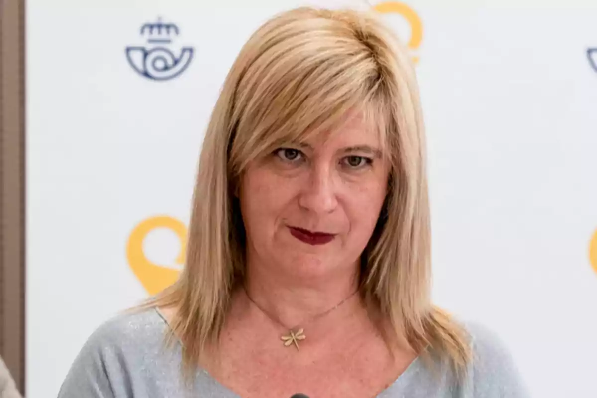 Mujer rubia de cabello lacio y suelto con expresión seria, lleva un collar con dije de libélula y está frente a un fondo blanco con logotipos amarillos y azules. Mujer rubia de cabello lacio y suelto con expresión seria, lleva un collar con dije de libélula y está frente a un fondo blanco con logotipos amarillos y azules.