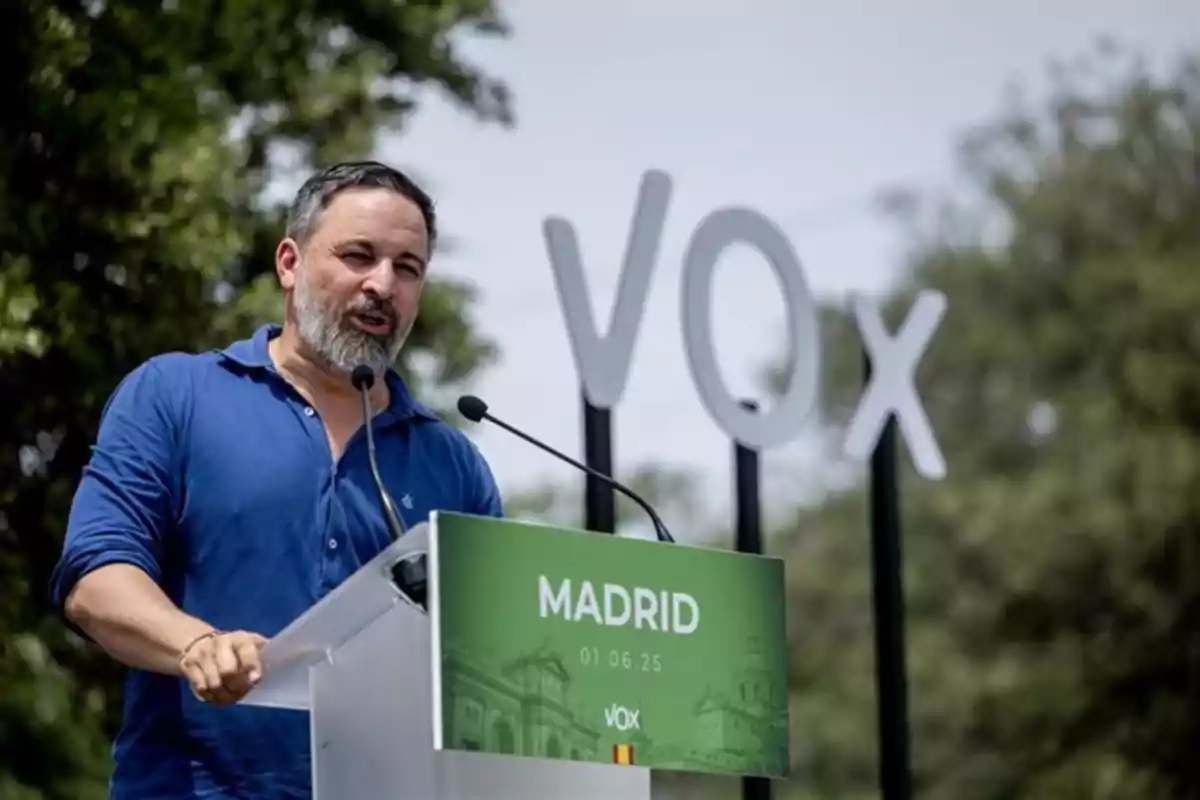 Hombre con barba y camisa azul hablando en un atril con un cartel verde que dice Madrid y el logo de Vox al fondo en un evento al aire libre Hombre con barba y camisa azul hablando en un atril con un cartel verde que dice Madrid y el logo de Vox al fondo en un evento al aire libre