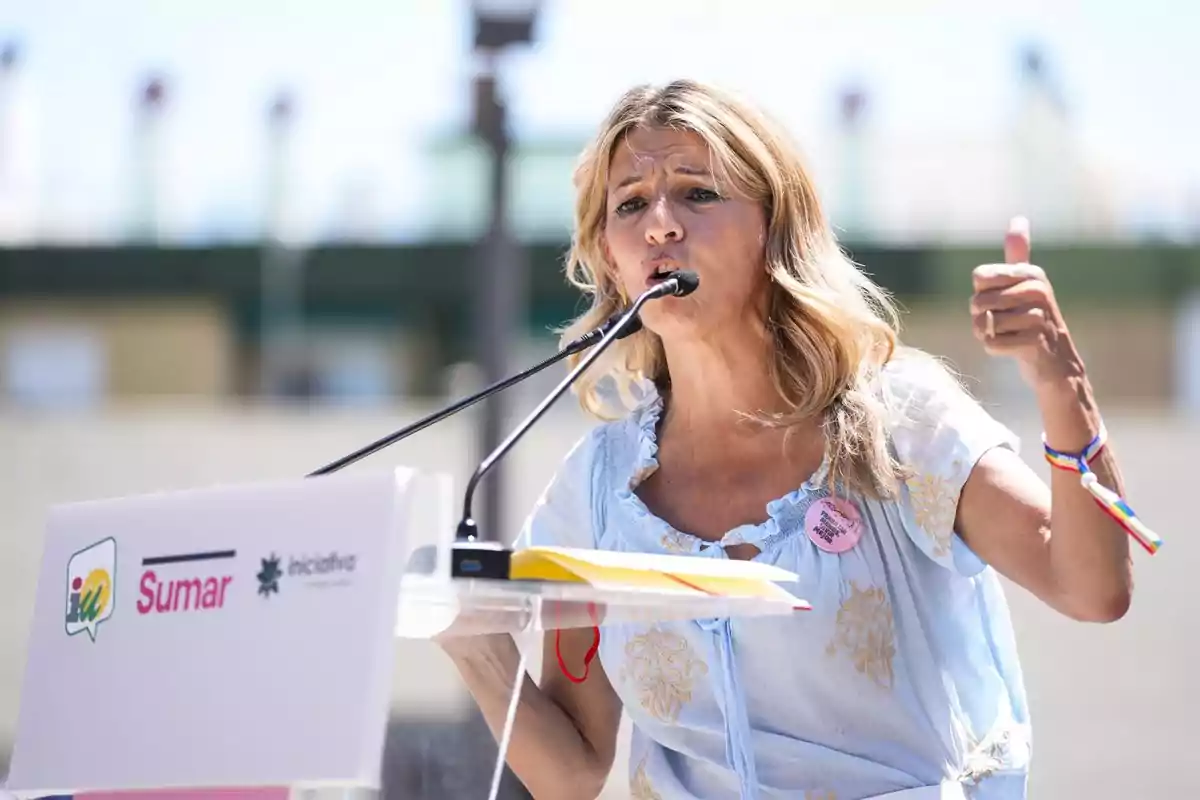 Mujer rubia hablando en un mitin al aire libre con micrófono y atril de la coalición Sumar, levantando el pulgar de la mano derecha y usando una pulsera de colores Mujer rubia hablando en un mitin al aire libre con micrófono y atril de la coalición Sumar, levantando el pulgar de la mano derecha y usando una pulsera de colores