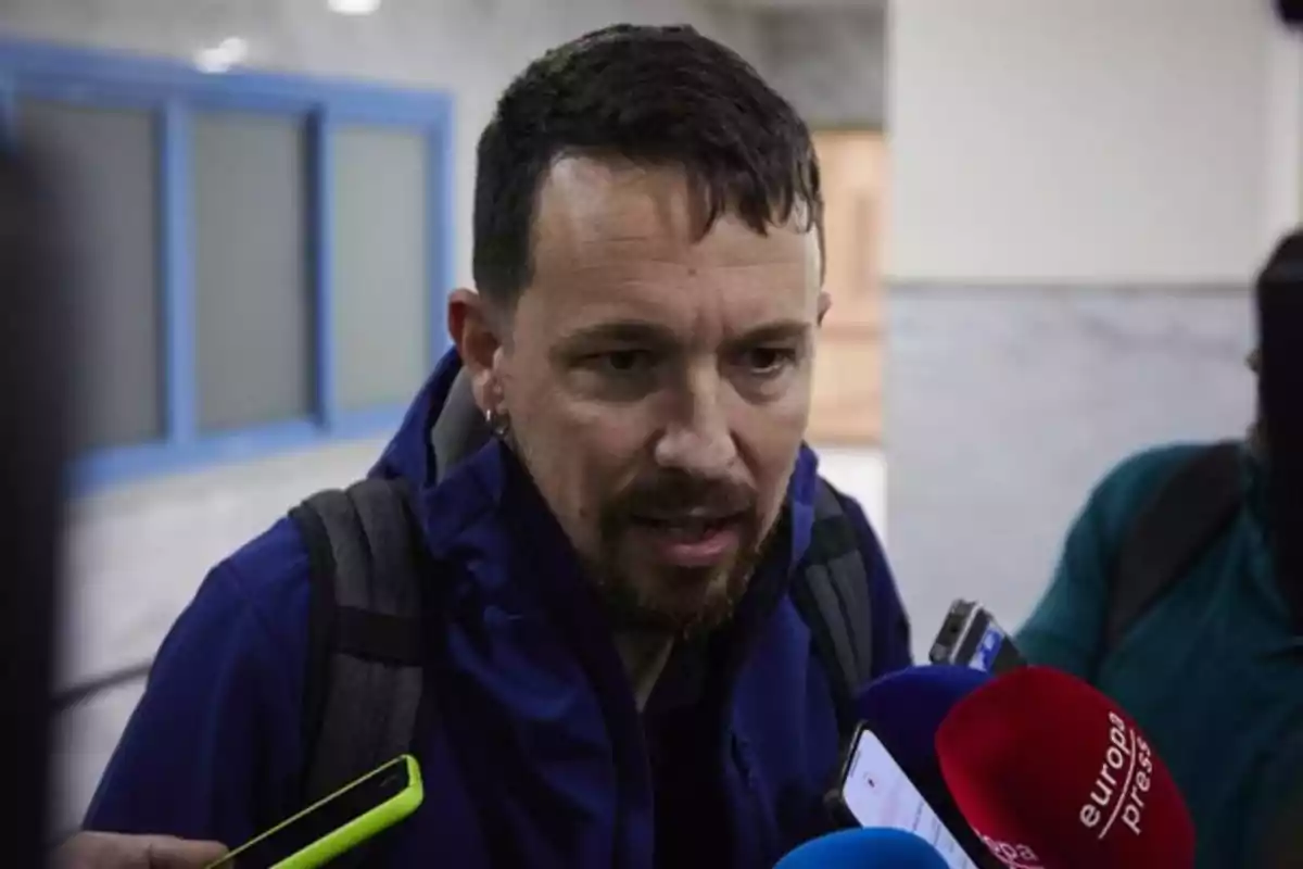 Hombre con barba y chaqueta azul responde preguntas de varios periodistas que sostienen micrófonos y grabadoras frente a él en un interior iluminado Hombre con barba y chaqueta azul responde preguntas de varios periodistas que sostienen micrófonos y grabadoras frente a él en un interior iluminado