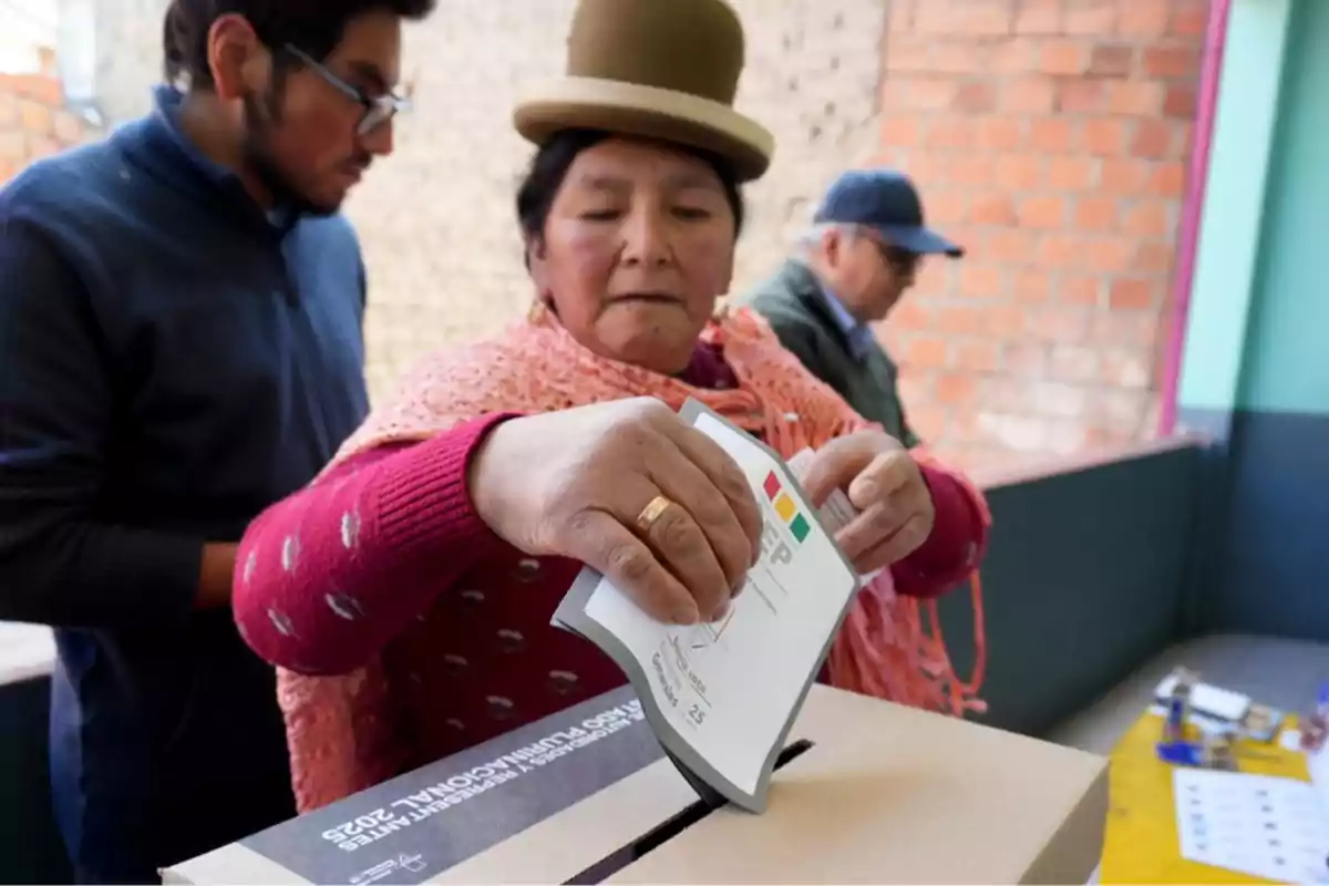 Mujer con sombrero tradicional deposita su voto en una urna durante una jornada electoral acompañada de otras personas Mujer con sombrero tradicional deposita su voto en una urna durante una jornada electoral acompañada de otras personas