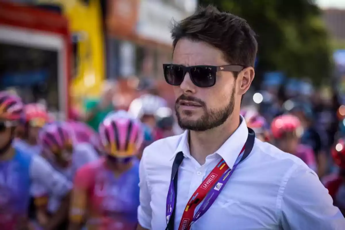 Hombre con gafas de sol y camisa blanca con gafete de La Vuelta al frente de un grupo de ciclistas con cascos coloridos