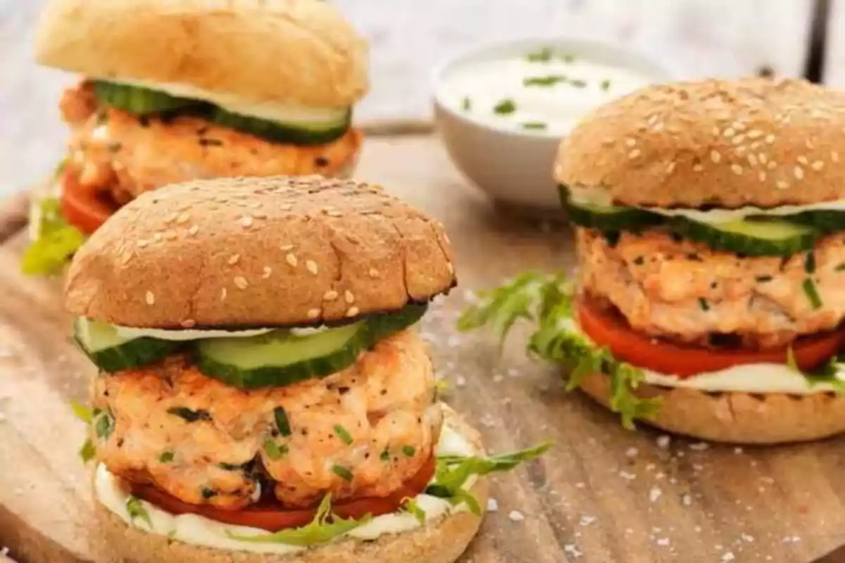 Tres hamburguesas de salmón con pepino, lechuga, tomate y mayonesa servidas en pan con ajonjolí sobre una tabla de madera