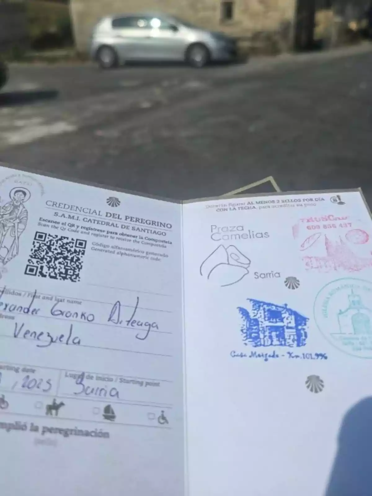 Una credencial del peregrino del Camino de Santiago con varios sellos y datos personales, sostenida al aire libre con un coche plateado y una calle al fondo Una credencial del peregrino del Camino de Santiago con varios sellos y datos personales, sostenida al aire libre con un coche plateado y una calle al fondo