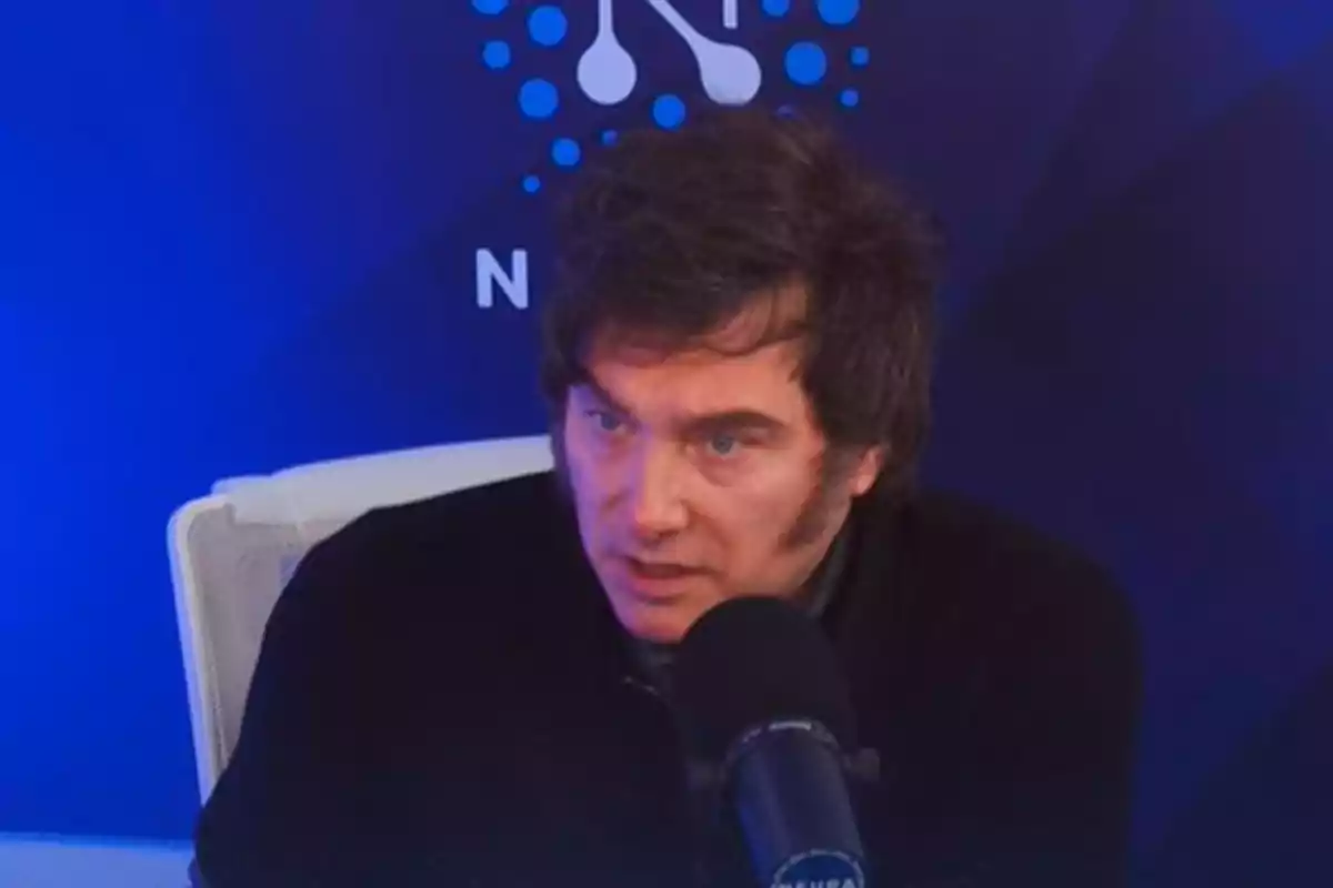 Hombre de cabello oscuro hablando frente a un micrófono en un estudio con fondo azul