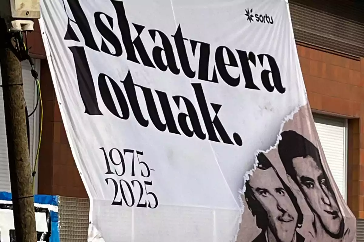 Pancarta blanca con letras negras que dice Askatzera lotuak 1975-2025, con el logo de Sortu en la esquina superior derecha y dos rostros dibujados en la parte inferior derecha. Pancarta blanca con letras negras que dice Askatzera lotuak 1975-2025, con el logo de Sortu en la esquina superior derecha y dos rostros dibujados en la parte inferior derecha.