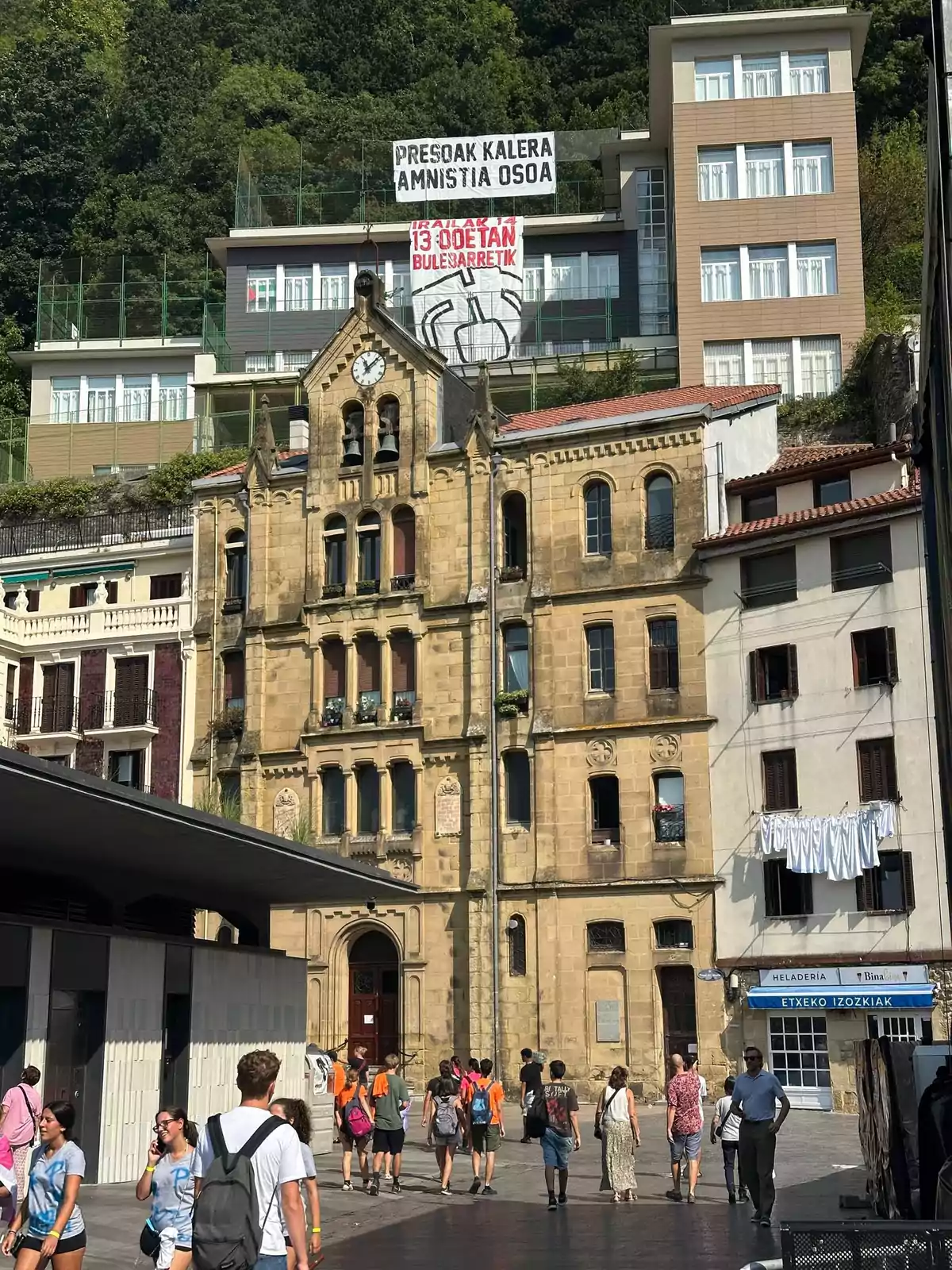 Personas caminando por una plaza frente a un edificio antiguo de piedra con un reloj en la parte superior y pancartas colgadas en los edificios de fondo Personas caminando por una plaza frente a un edificio antiguo de piedra con un reloj en la parte superior y pancartas colgadas en los edificios de fondo