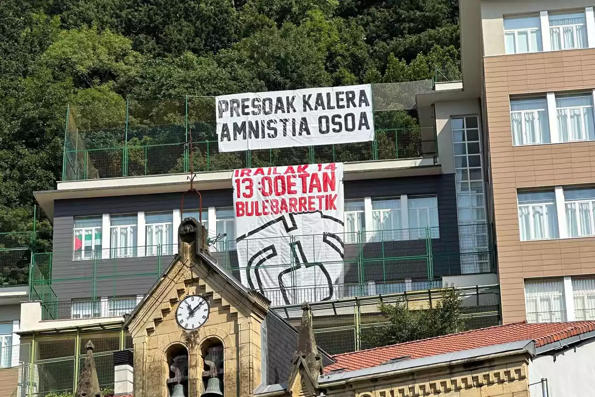 Dos pancartas con mensajes en euskera cuelgan en la fachada de un edificio moderno detrás de una iglesia con un reloj y campanario, rodeado de vegetación y árboles. Dos pancartas con mensajes en euskera cuelgan en la fachada de un edificio moderno detrás de una iglesia con un reloj y campanario, rodeado de vegetación y árboles.