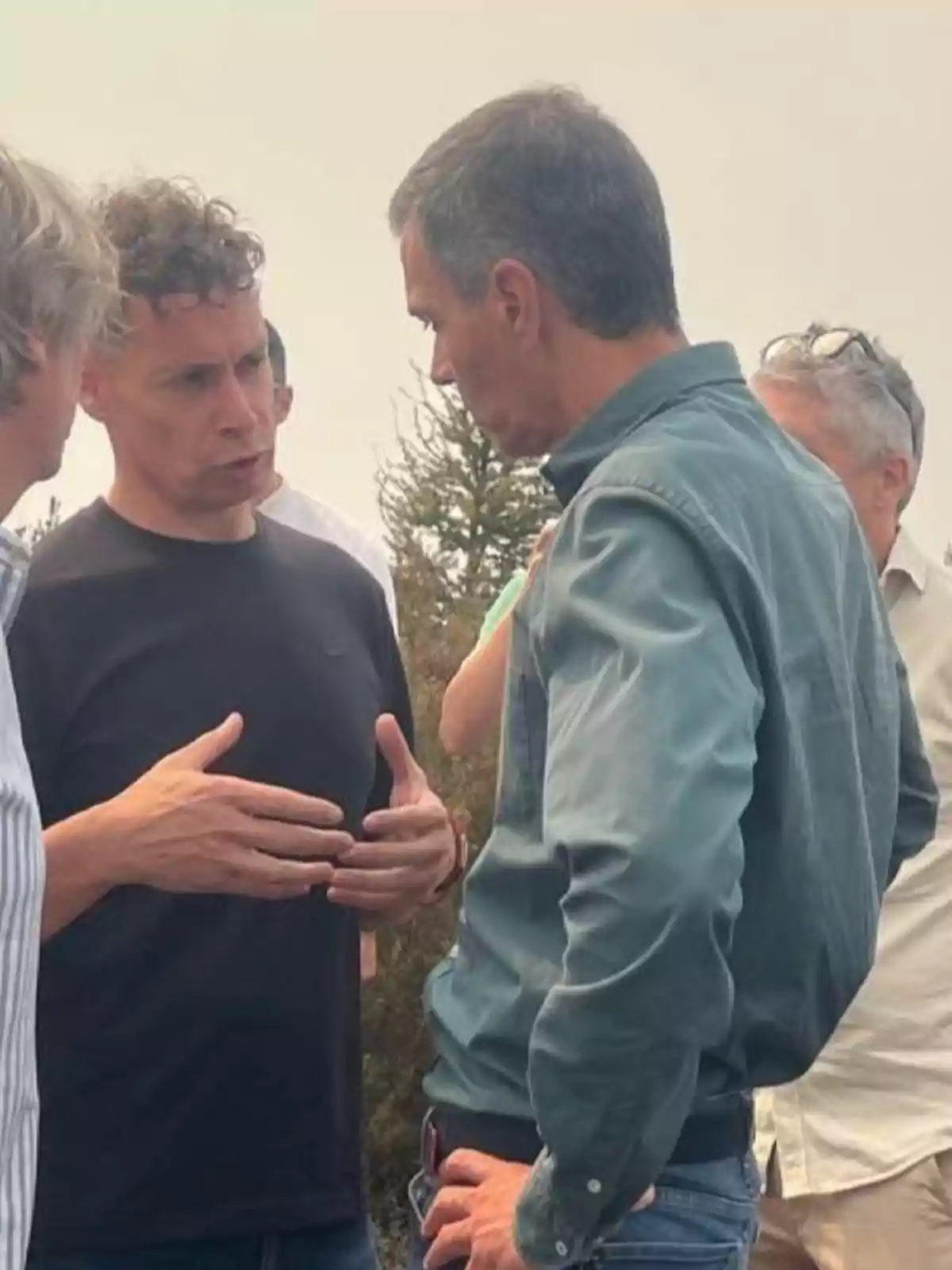 Dos hombres conversan de manera seria al aire libre mientras otras personas están alrededor Dos hombres conversan de manera seria al aire libre mientras otras personas están alrededor